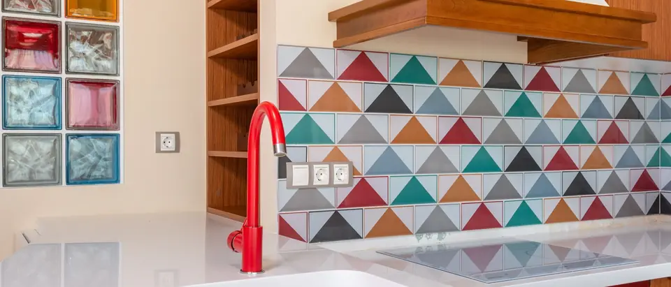 A bright kitchen with a red faucet over a white sink and a colorful tiled wall