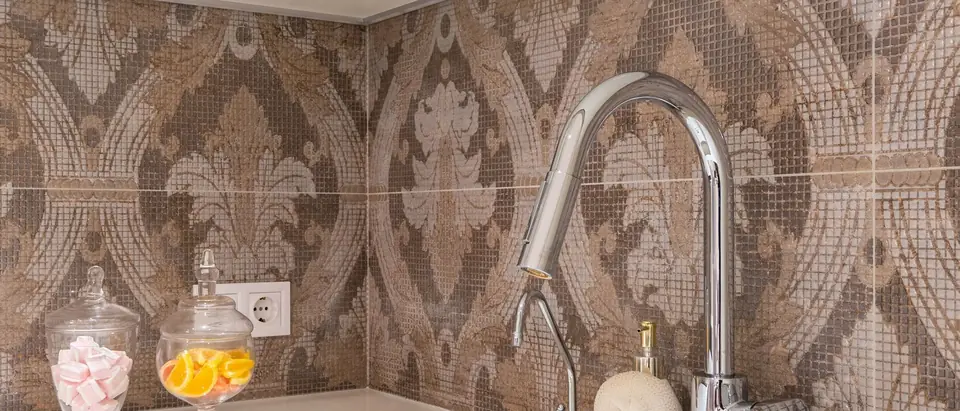A chrome kitchen faucet and a white sink against a patterned tile wall