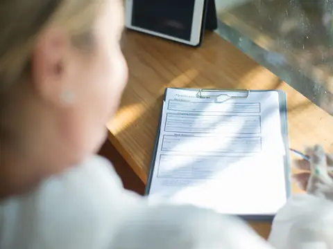 A hand filling in a form with a pen at a sunlit wooden desk