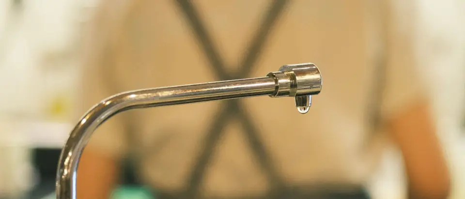 A drop of water hanging from the spout of a metal faucet
