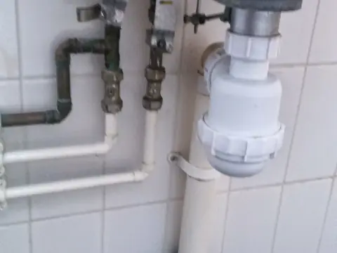 Drain pipes and supply lines under a sink against white tiles